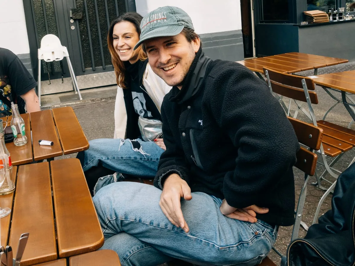 Outside a bar: three people at pushed-together wooden tables, one turning to the camera laughing