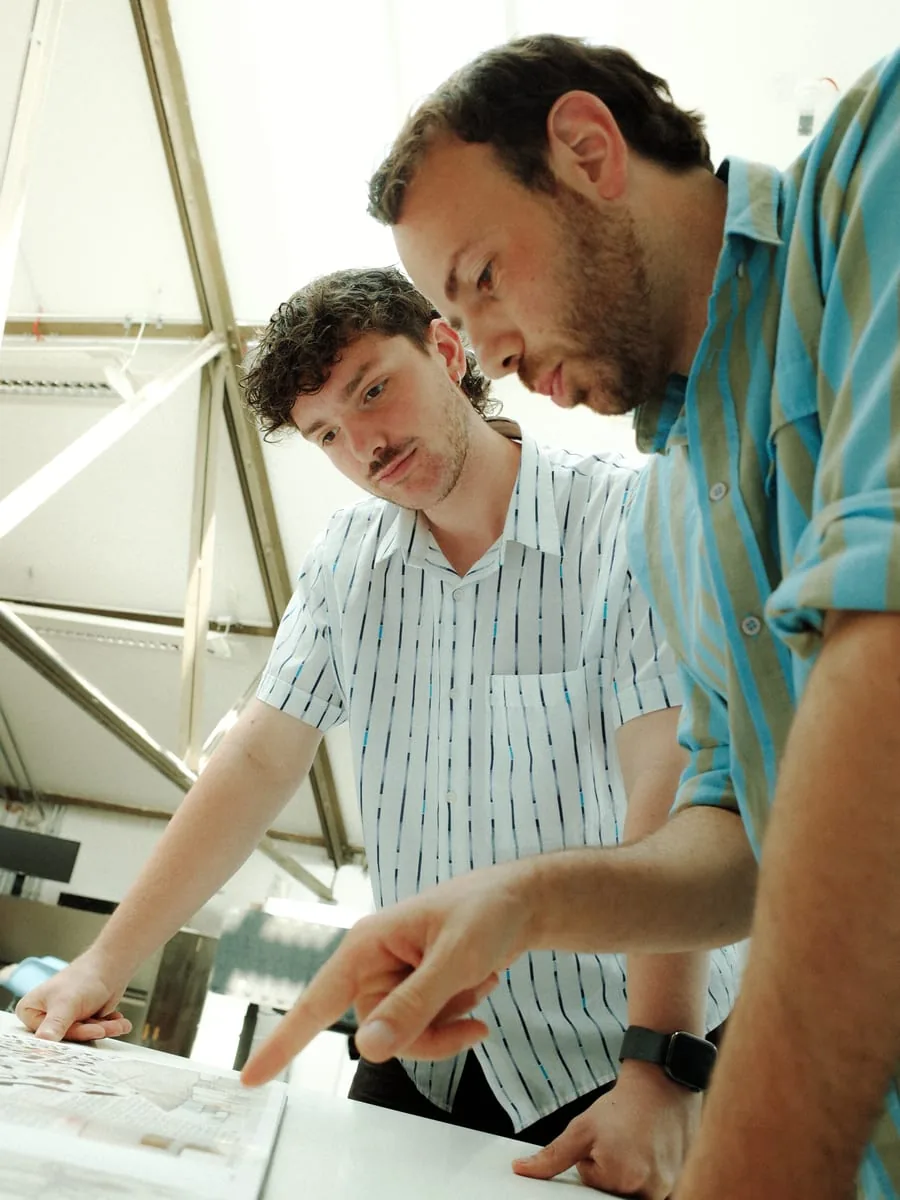 Concept work before the content creation: two men standing over a storyboard laid out flat, one pointing at a frame, the roof structure of the hall behind them