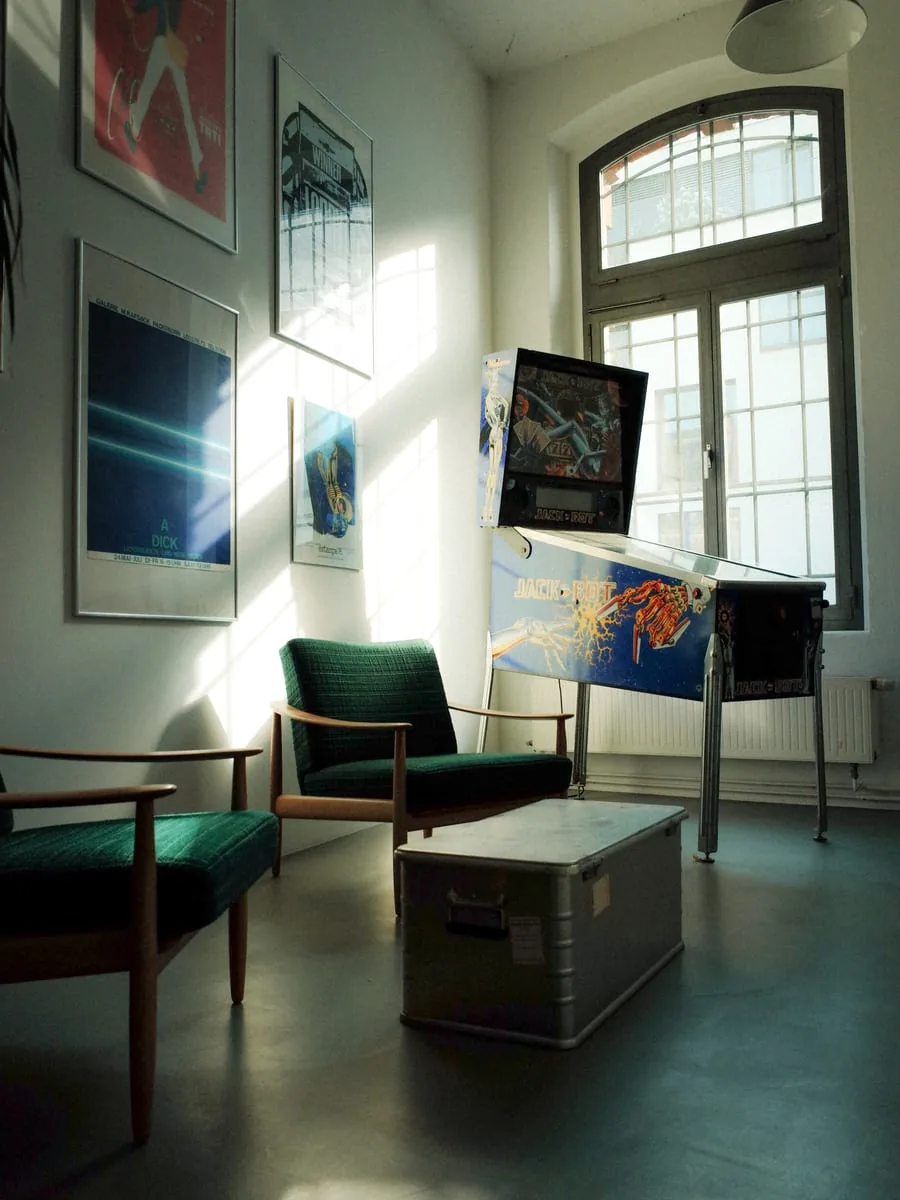 A corner of the Krafthaus office in Cologne: two green armchairs and a pinball machine below a tall arched window, posters on the wall