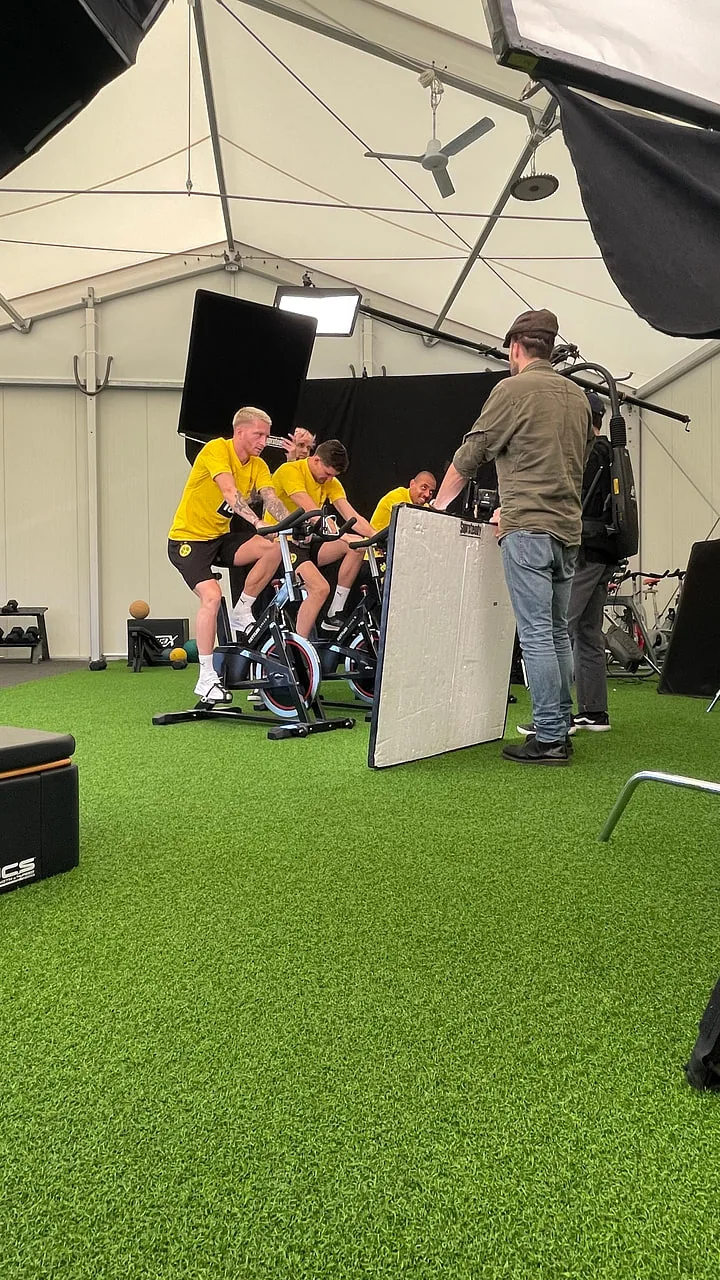 Social Ad für BVB × EA Sports: Dreh auf den Spinning-Rädern Aus der Produktion der Social Ad: die Spieler auf den Spinning-Rädern, davor die Crew mit einem Reflektor, dahinter die helle Wand des Zelts