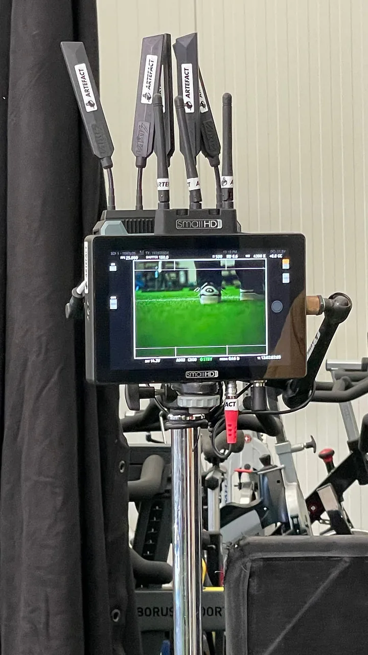 Regiemonitor mit dem Kunstrasen aus dem Spiel Regiemonitor mit Funkantennen auf einem Stativ zeigt den Kunstrasen, dahinter die Spinning-Räder und ein schwarzer Vorhang