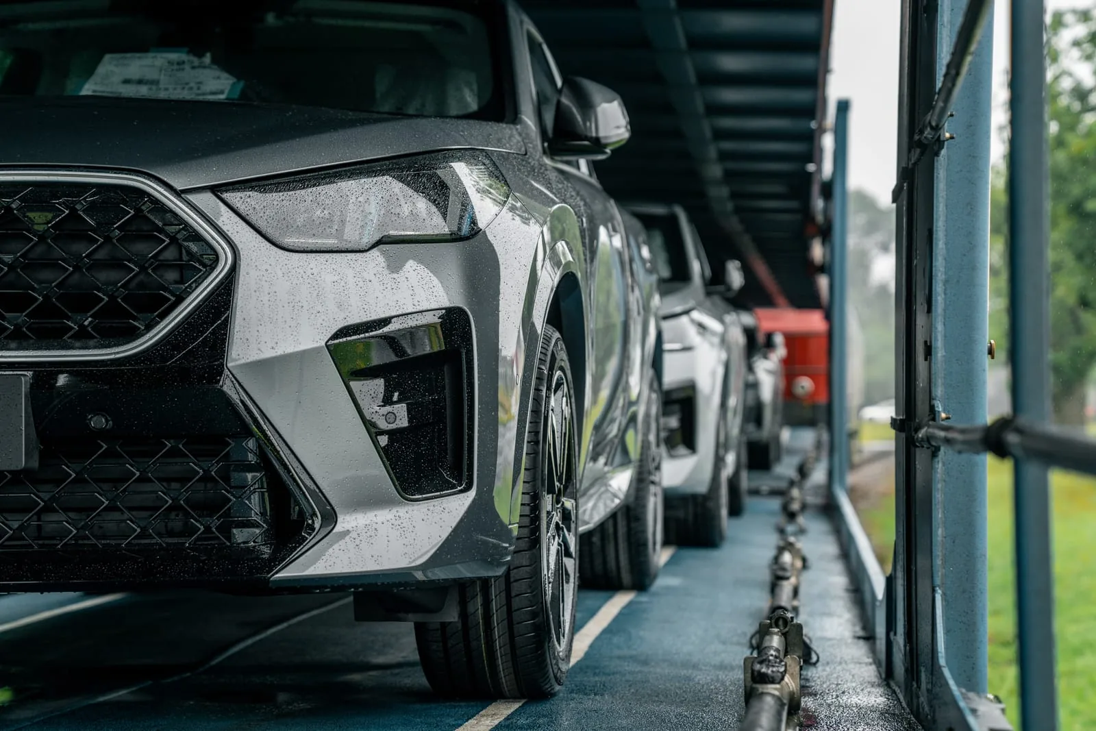 Fahrzeug auf dem Autotransporter Regennasser grauer SUV auf dem Deck eines Autotransporters, dahinter weitere Wagen, rechts das Grün am Rand