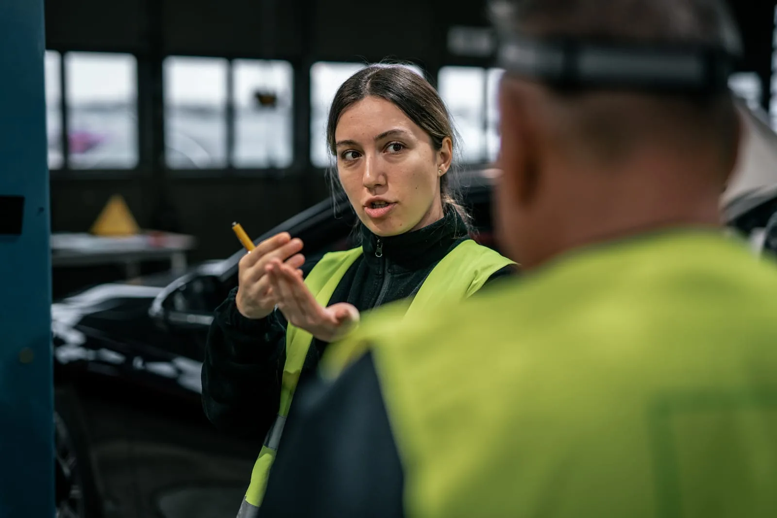 Mitarbeitende im Gespräch in der Werkhalle Frau in Warnweste erklärt mit einem Stift in der Hand etwas, ihr Gegenüber im Anschnitt, dahinter ein dunkles Fahrzeug