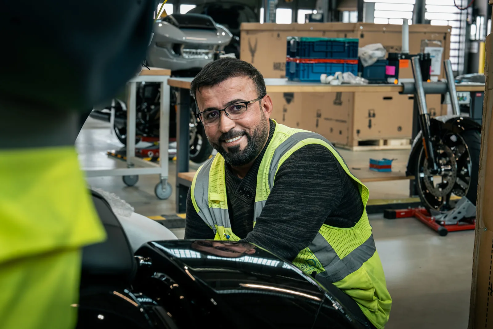 Mitarbeiterporträt in der Werkstatt von Formel D Mann mit Brille in Warnweste lächelt über eine schwarze Motorradverkleidung hinweg in die Kamera