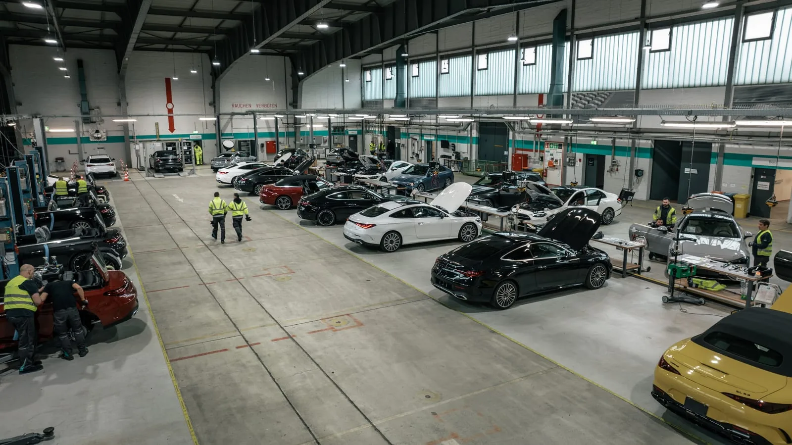 Überblick über die Werkhalle von Formel D Unternehmensfotografie vor Ort: Überblick über die Halle mit Fahrzeugen an Hebebühnen und Werkbänken, dazwischen Mitarbeitende in Warnwesten
