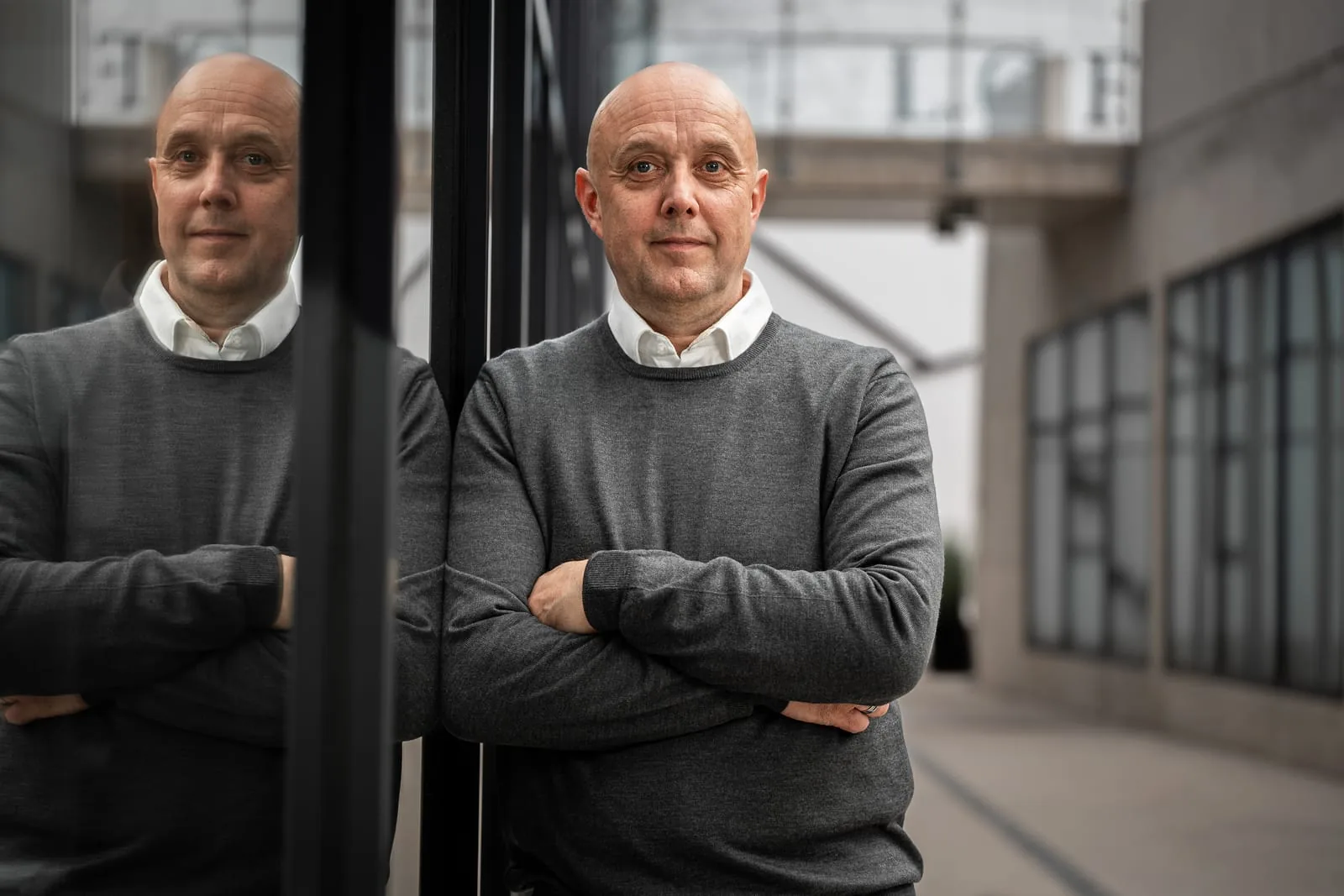 Unternehmensporträt an der Glasfassade Mann im grauen Pullover lehnt mit verschränkten Armen an einer Glasscheibe, die ihn spiegelt, dahinter der Innenhof