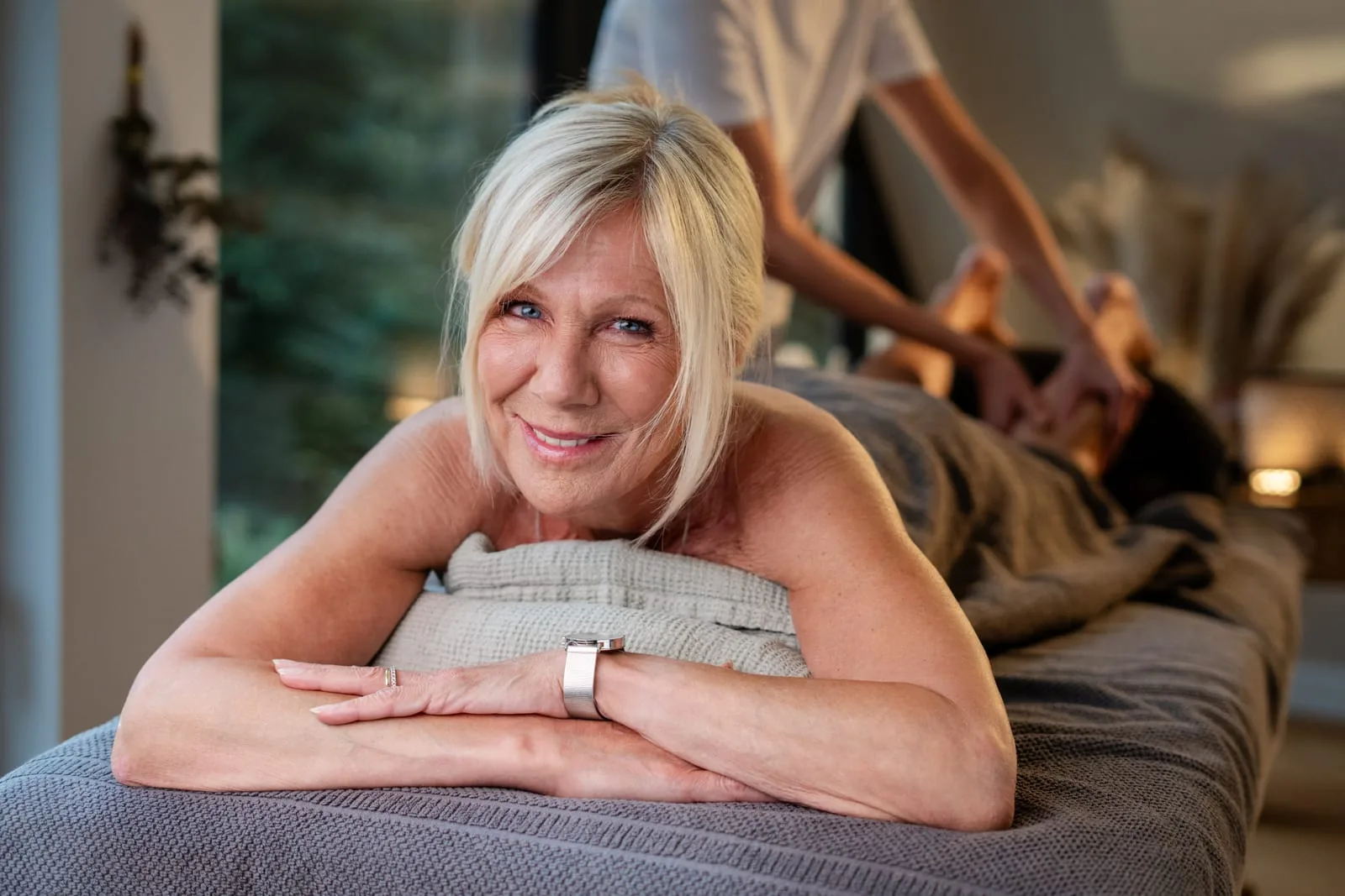 Frau liegt lächelnd auf der Massageliege, im Hintergrund massieren zwei Hände ihre Wade