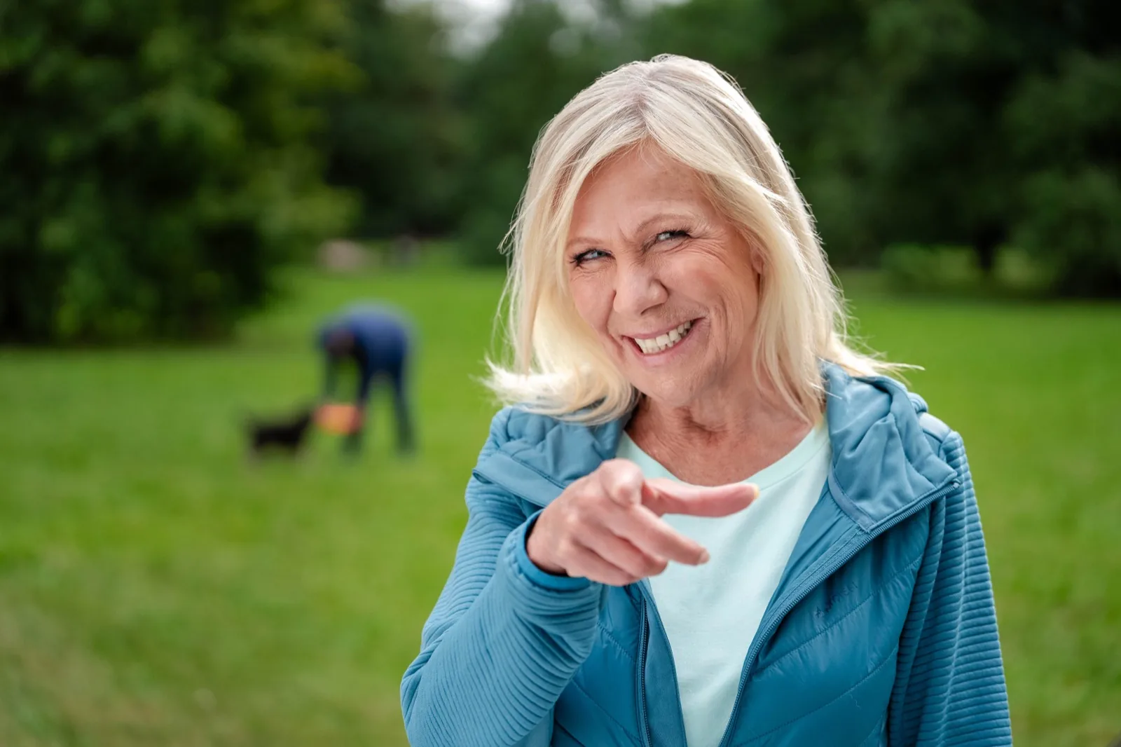 Ulrike von der Groeben zeigt lachend in die Kamera, hinter ihr bückt sich jemand im Park zu einem kleinen Hund