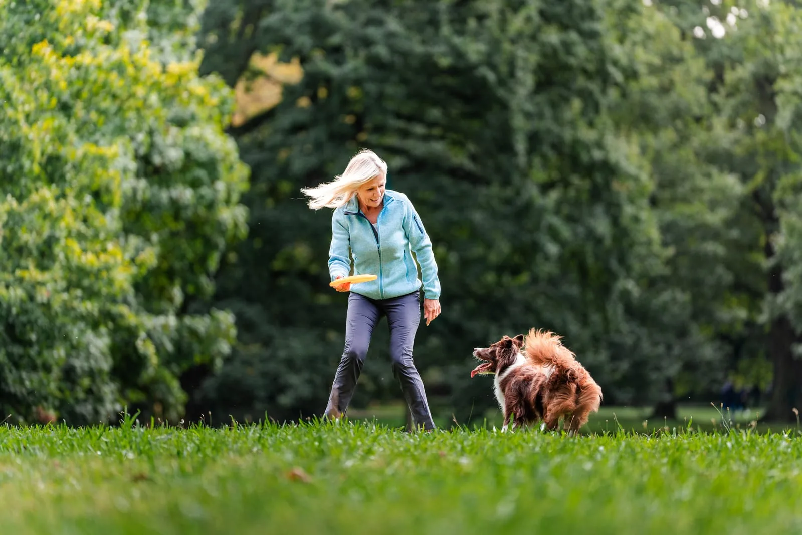 Aus der Testimonial-Kampagne mit Ulrike von der Groeben: sie hält im Park eine orangefarbene Wurfscheibe, neben ihr ein brauner Border Collie