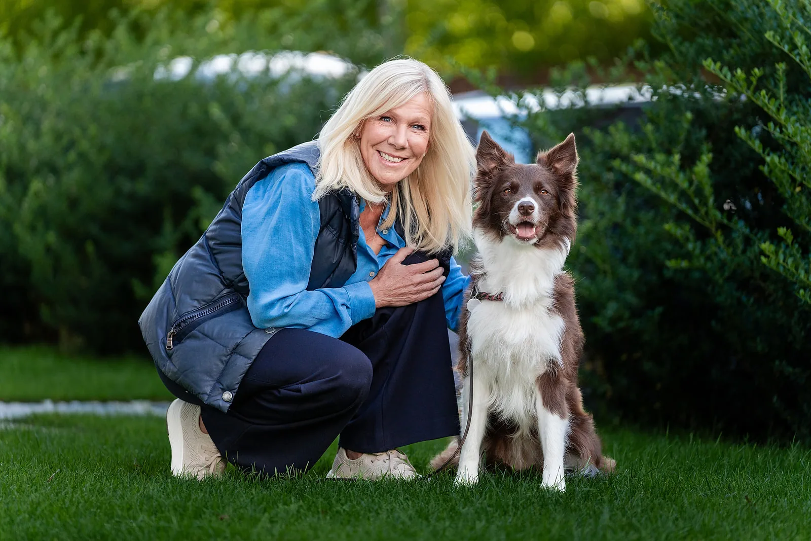 Ulrike von der Groeben hockt lächelnd neben einem braun-weißen Border Collie im Gras, die Hand auf der Brust