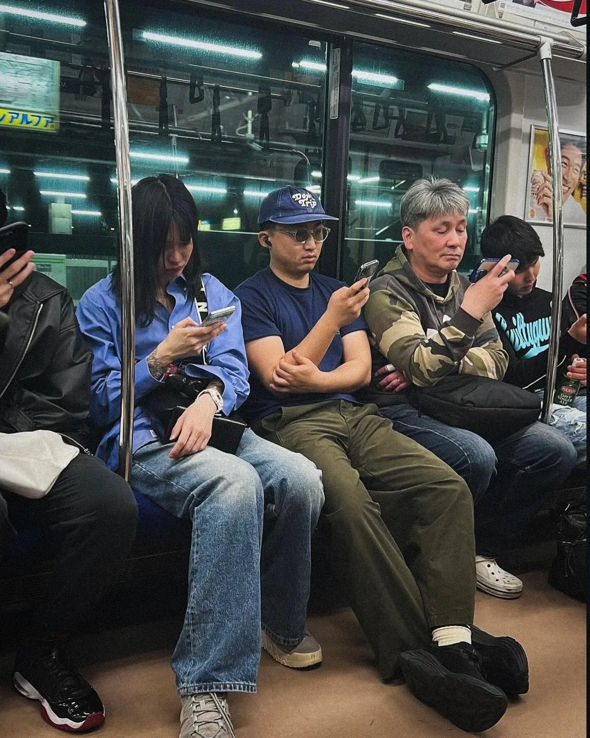 The audience for ongoing social media content: a row of seats on the underground, six people side by side, nearly all of them looking at a phone