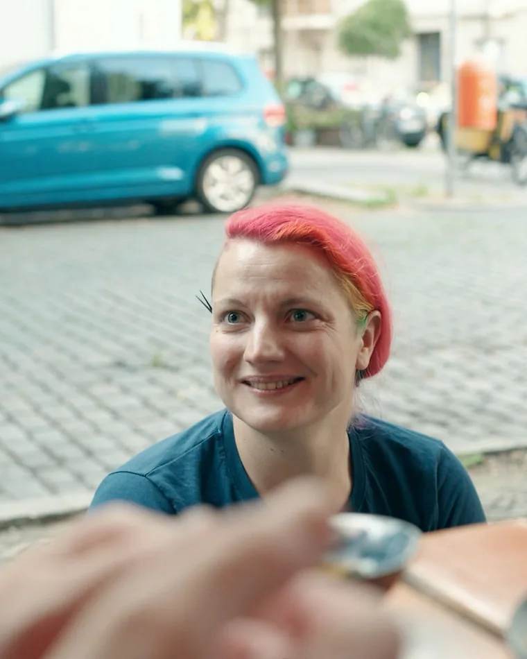Mitarbeiterporträt für Siemens: Lydia Lydia aus dem Mitarbeiterporträt für Siemens: pink gefärbtes Haar, sie lächelt am Tisch vorbei an der Kamera, dahinter Kopfsteinpflaster und ein blauer Van