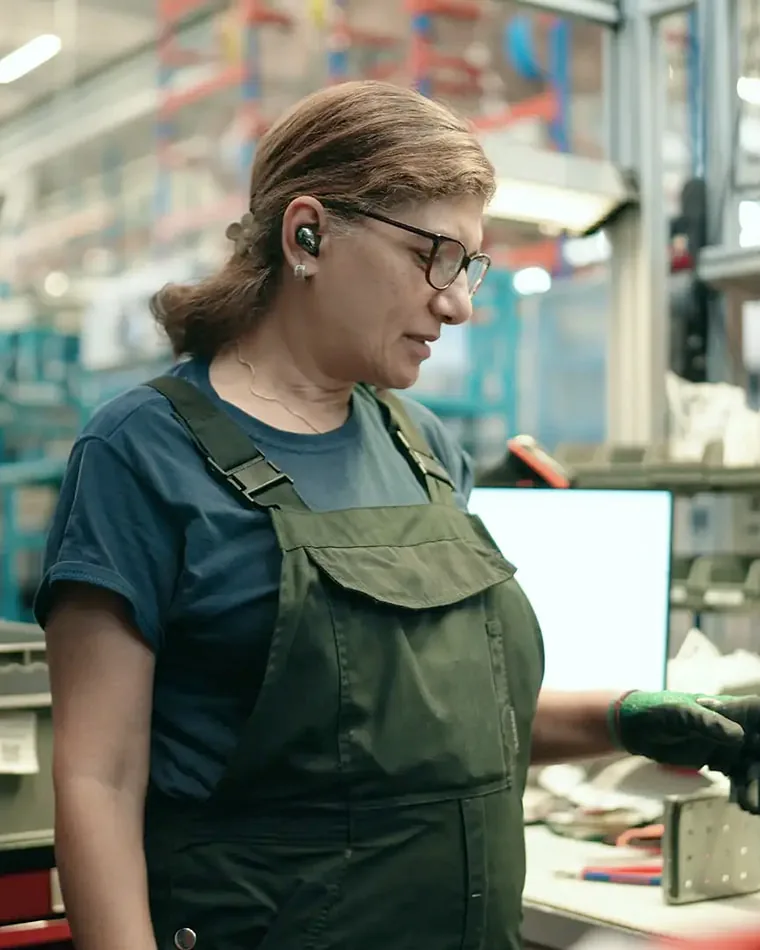 Mitarbeiterin an der Werkbank in der Fertigung Eine Mitarbeiterin in Latzhose prüft an der Werkbank ein Bauteil, hinter ihr die Fertigung