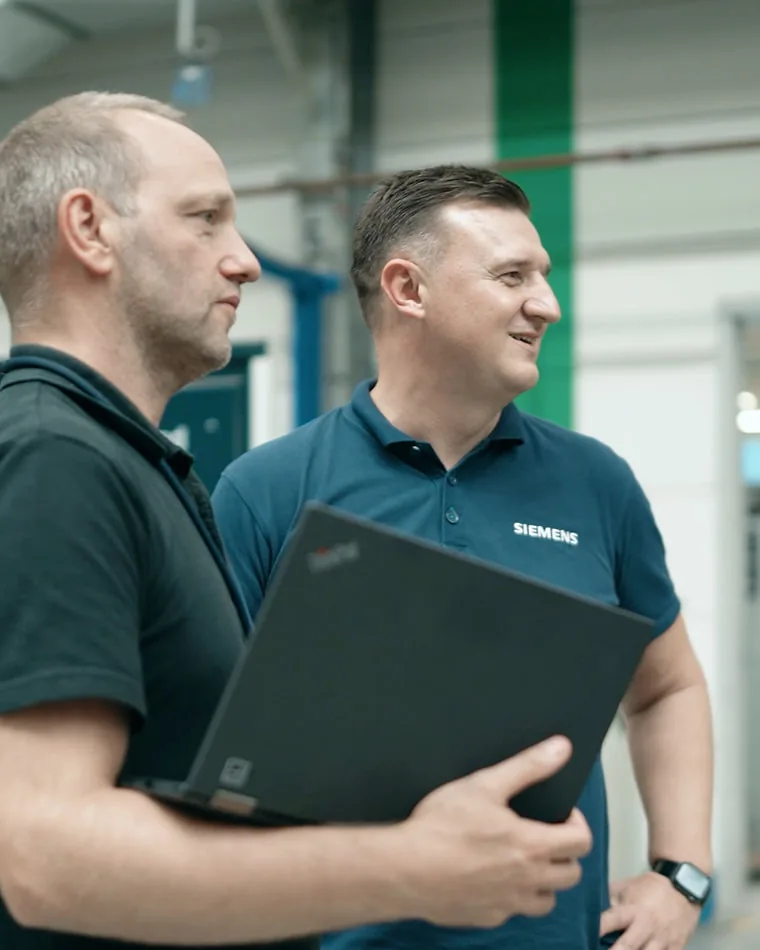Vedad im Gespräch mit einem Kollegen Vedad im Polohemd mit SIEMENS-Schriftzug geht lachend neben einem Kollegen, der einen Laptop unter dem Arm trägt