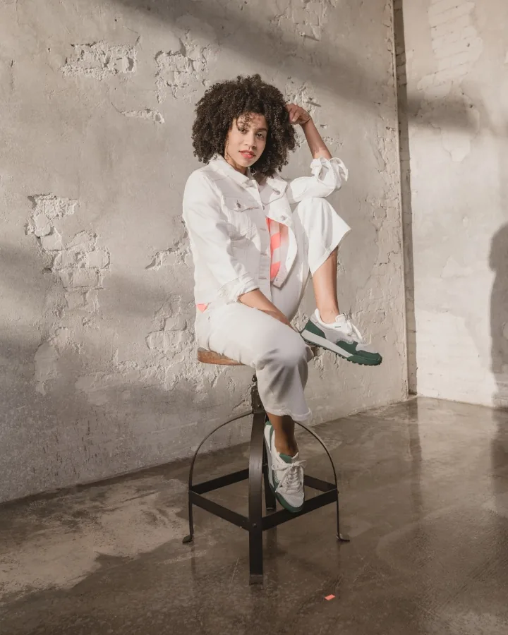 Final look of the series for the fashion brand: model dressed all in white on a black stool, one leg drawn up, on the polished concrete floor