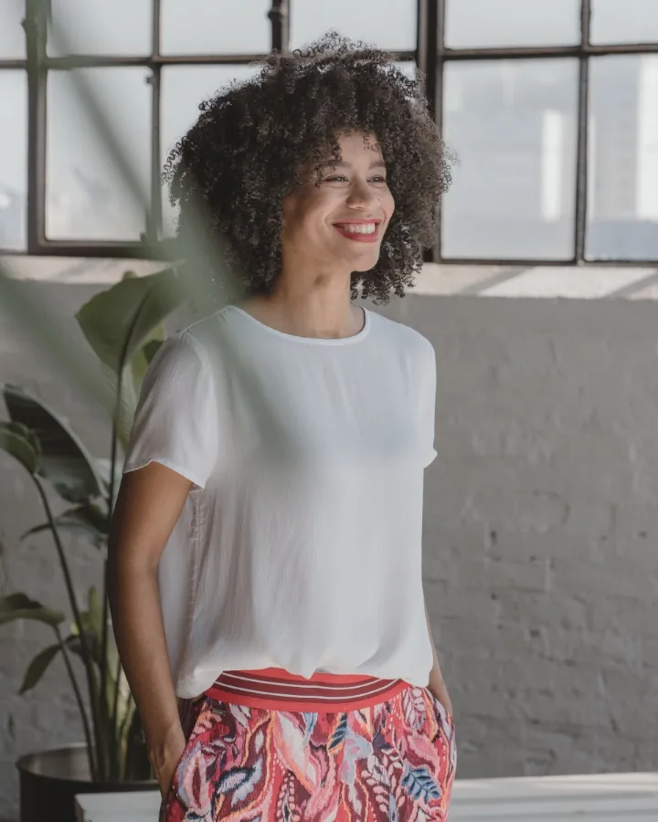 Model in a white shirt and red patterned trousers laughing by a paned window, a large plant at left
