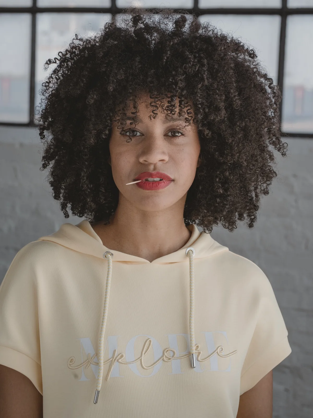 Social content for the fashion brand Street One: model in a pale hoodie reading “explore MORE”, a toothpick at the corner of her mouth, the window behind