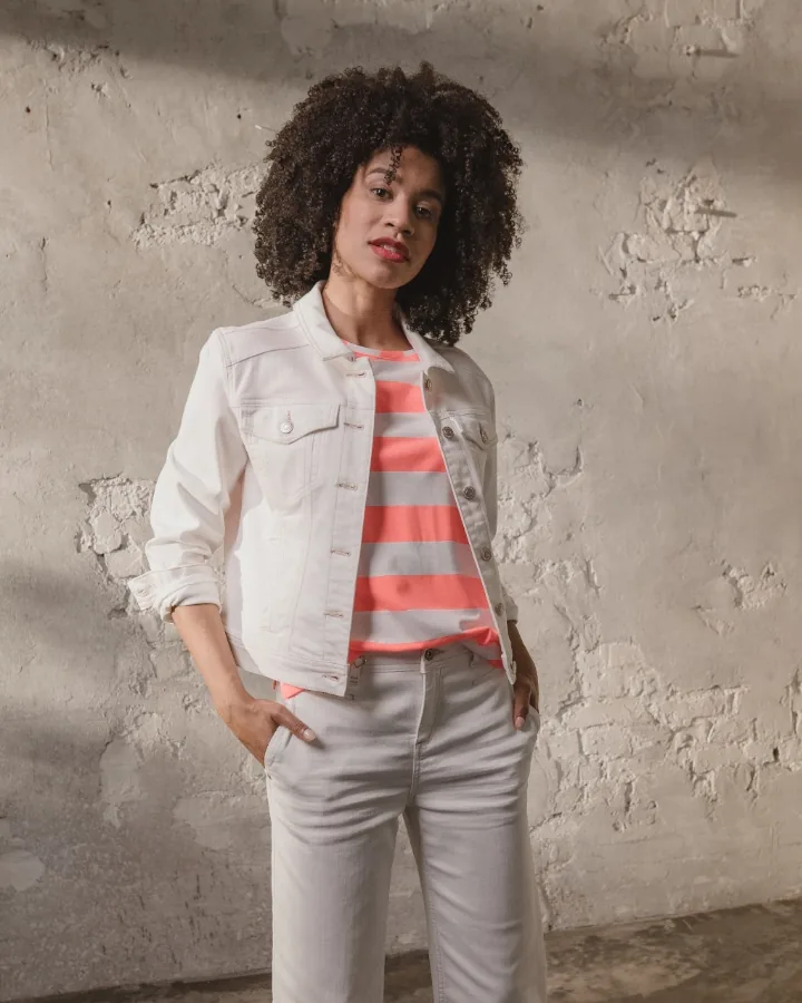 Model in a white denim jacket over a coral and grey striped shirt and pale trousers by a rough rendered wall