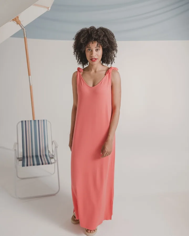 Summer look for the fashion brand: model in a long coral dress beside a parasol and a striped beach chair against a pale ground