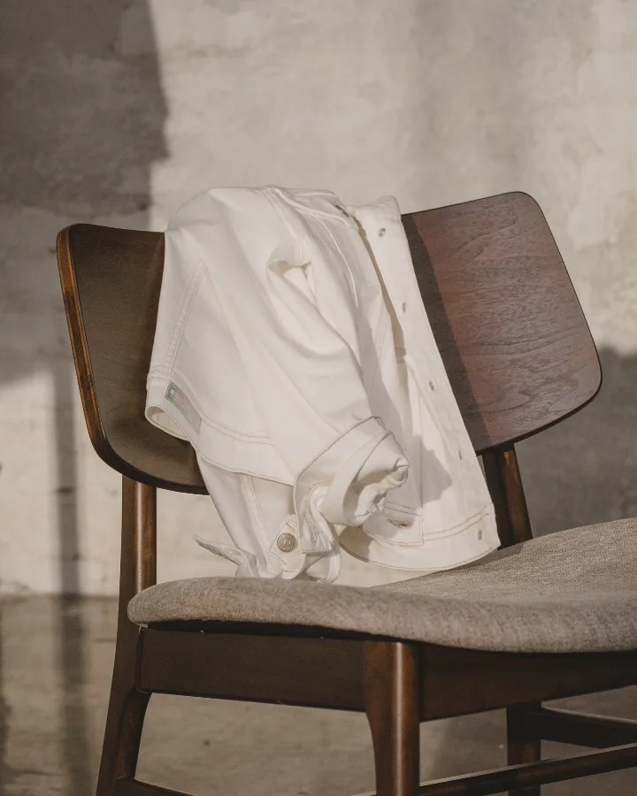 A white denim jacket over the back of a dark wooden chair, sunlight falling on the wall beside it