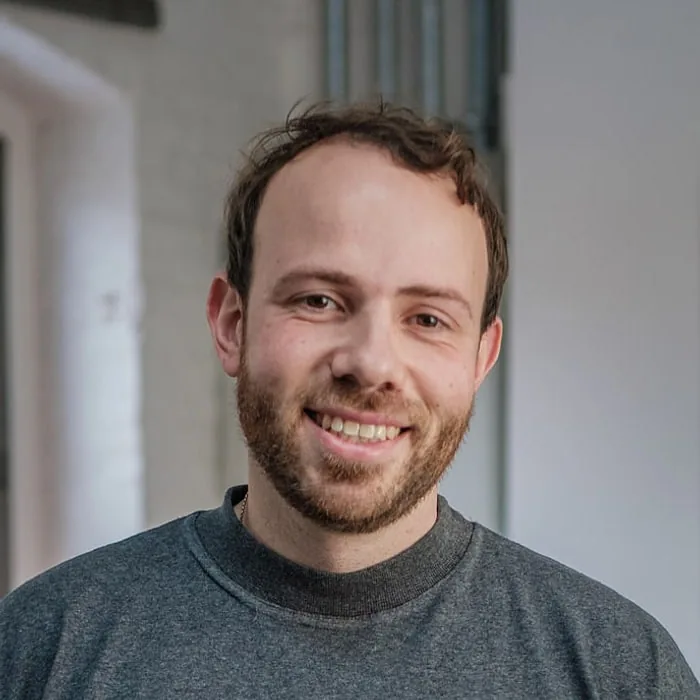 Cem-Pierre Schuch, head of production at Le Werk in Cologne, in a grey shirt, laughing in front of a white wall