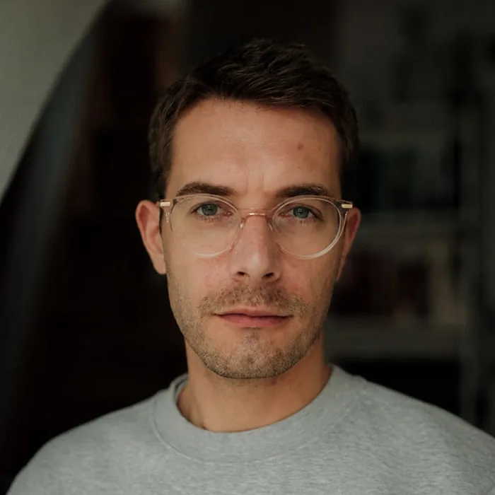 Joscha Ortmeier, head of post-production at Le Werk in Cologne, in light-framed glasses and a grey sweatshirt in front of a bookshelf