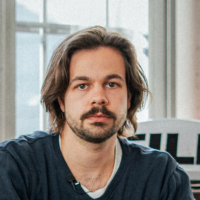 Tom Beckers, producer and content creator at Le Werk in Cologne, with shoulder-length hair and a moustache in front of a bright window