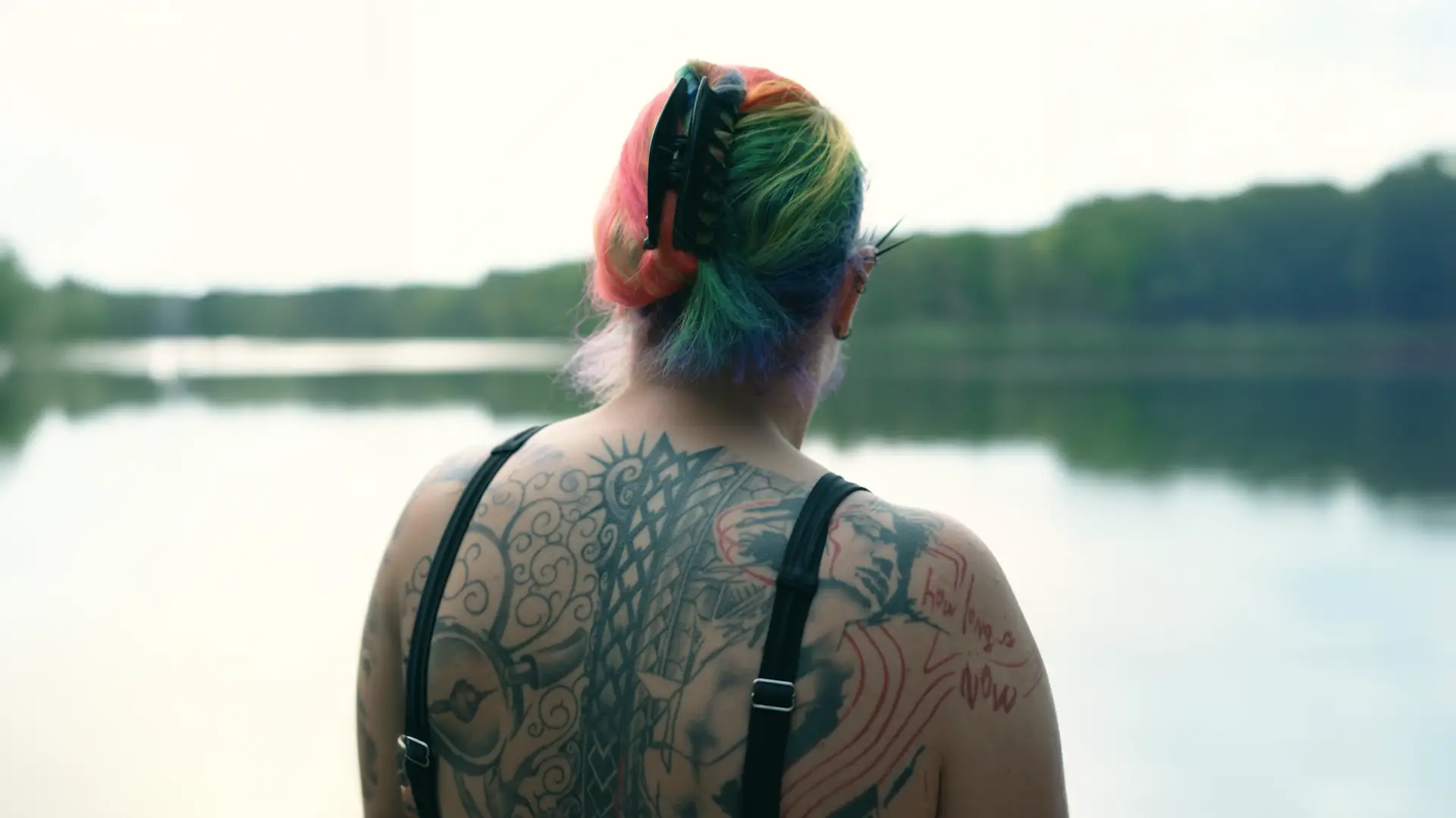 Still from an employee portrait for Siemens: woman with rainbow-dyed hair and a large back tattoo at the edge of a lake, seen from behind