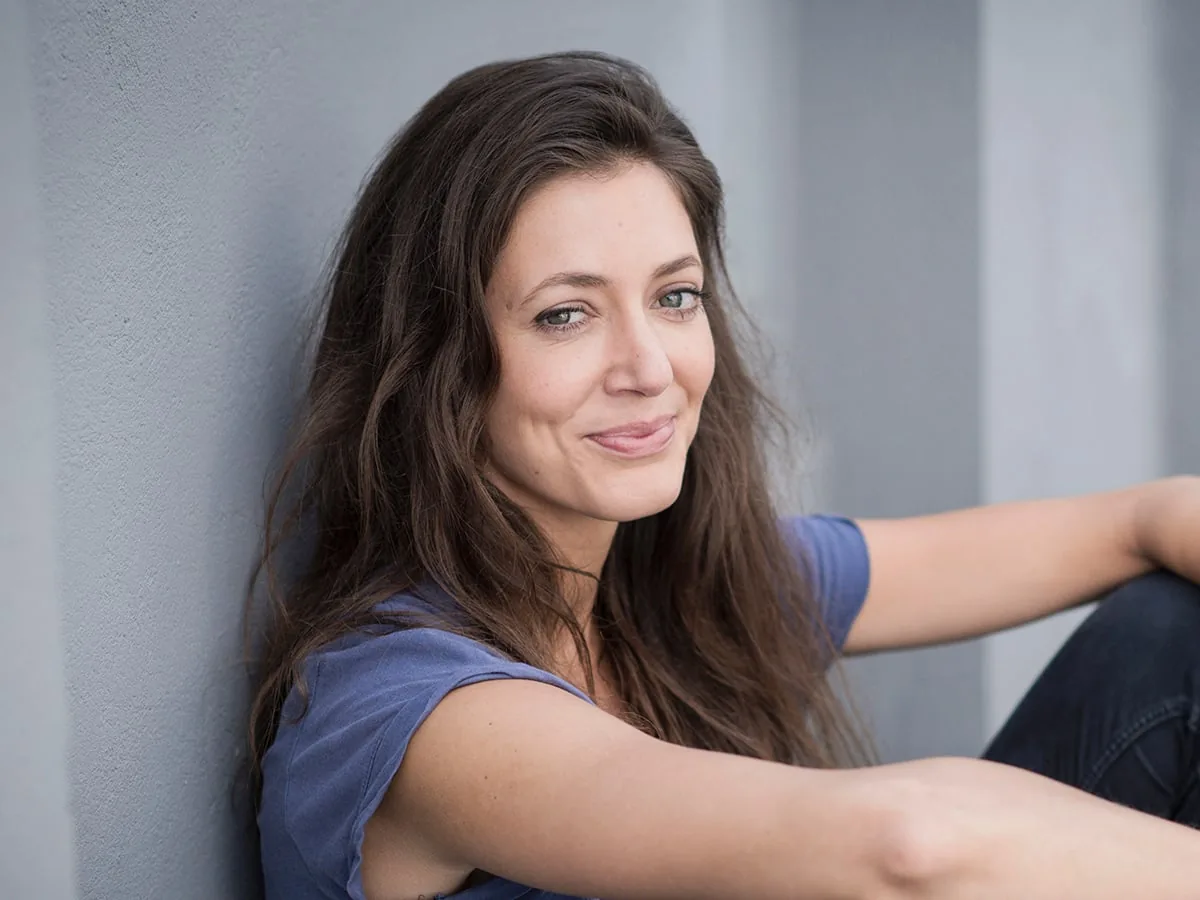 Anna Möbus — actor and coach Anna Möbus, actor and coach in the workshops: she sits leaning against a grey wall, one arm on her knee, smiling into the camera