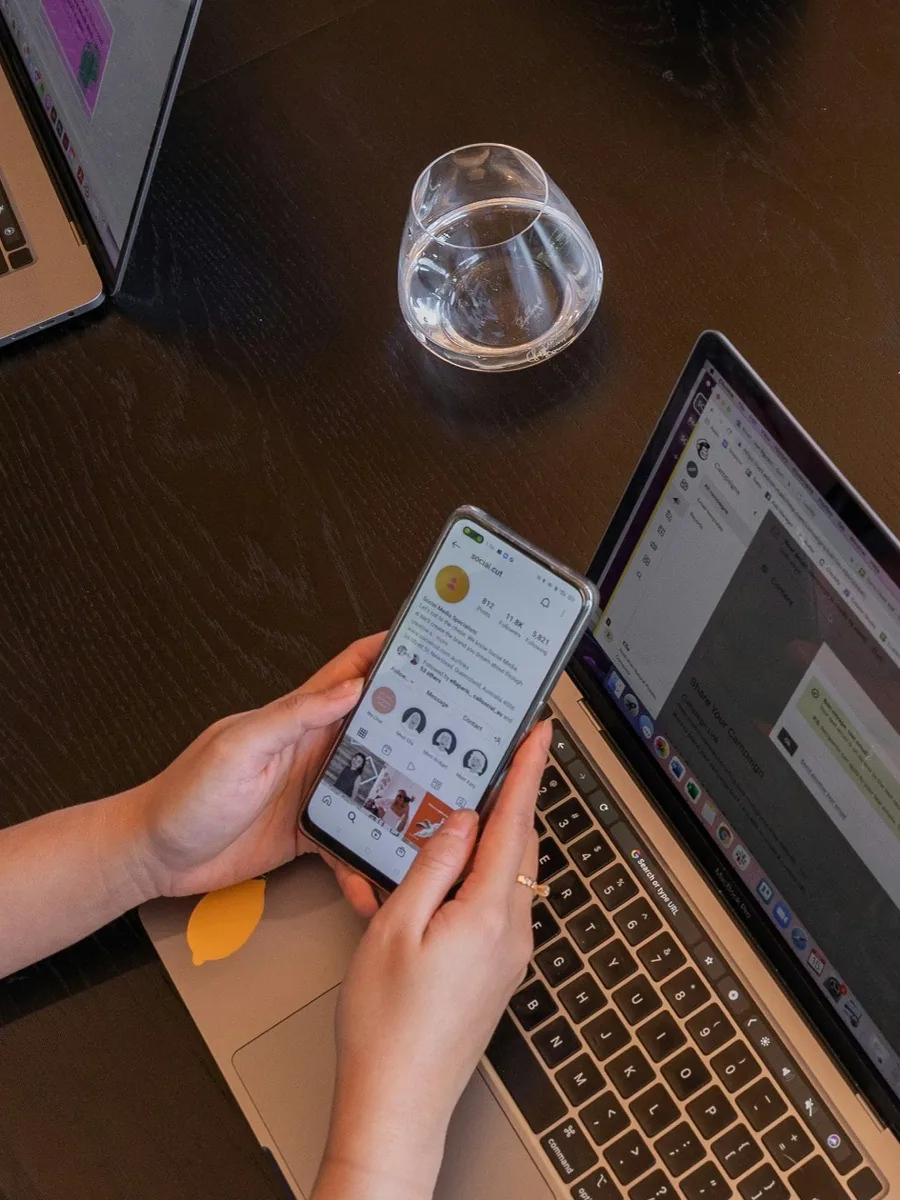 In the social media workshop: two hands holding a phone with a profile open above a dark table, an open laptop beside it