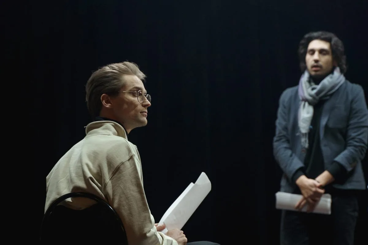 Scene from the improv workshop A scene from the improv workshop: two people on a dark stage, one sitting on a chair with a script, the other standing further back