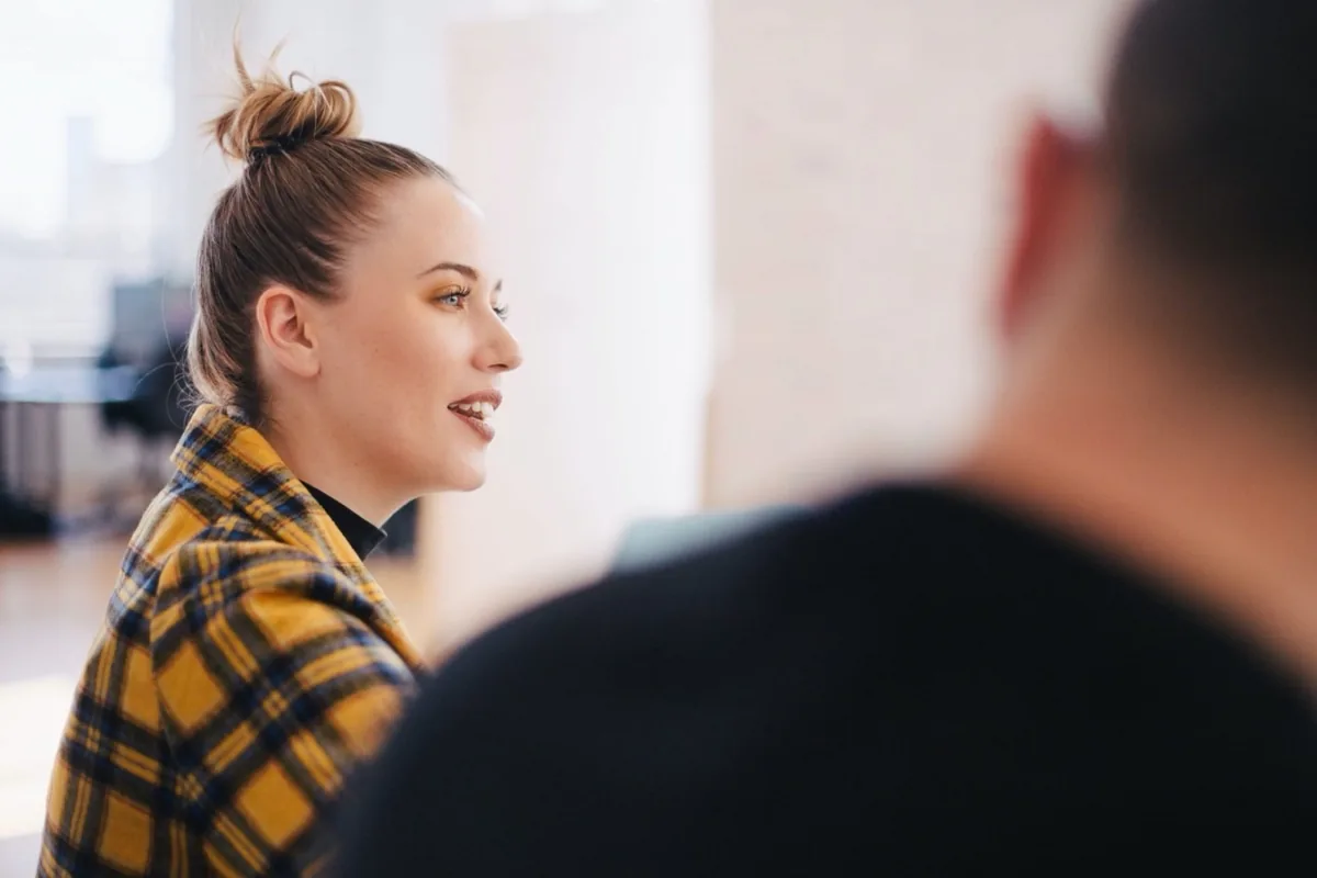 Exercise during the camera training Young woman in a yellow checked coat speaking in a bright room, the back of the person opposite blurred in the foreground