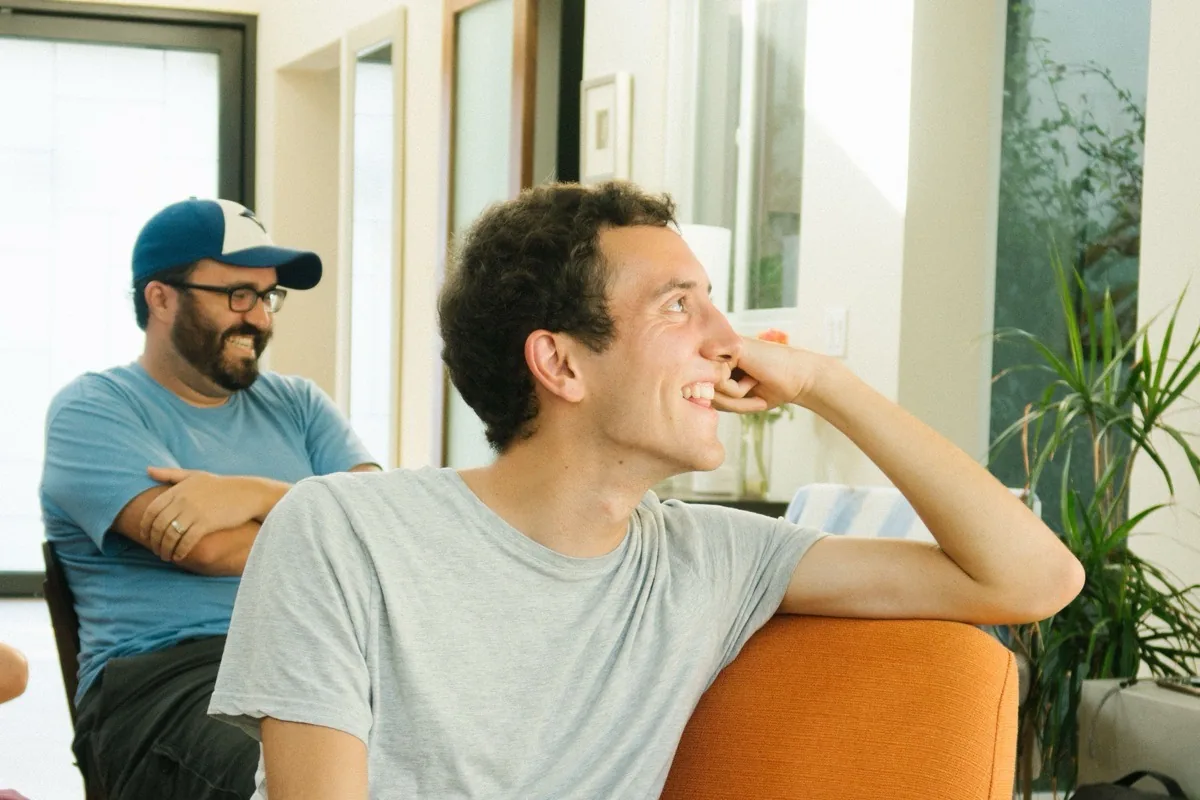 Listening as an exercise in improv Two men sitting in orange armchairs, listening; the one in front rests his chin in his hand, laughing